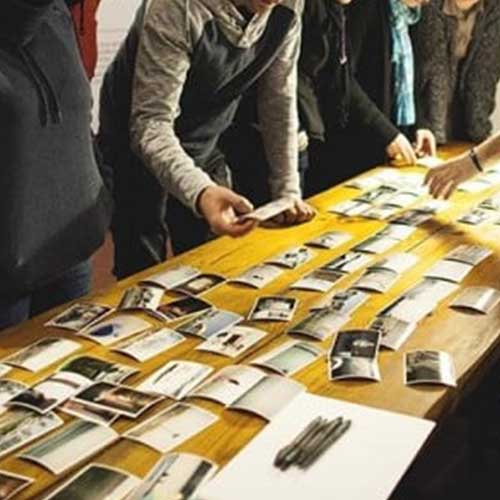 Na zdjęciu grupa osób stojąca przy stole podczas wyboru zdjęć, które ułożone są na stole. Nie widoczne są twarze uczestników.