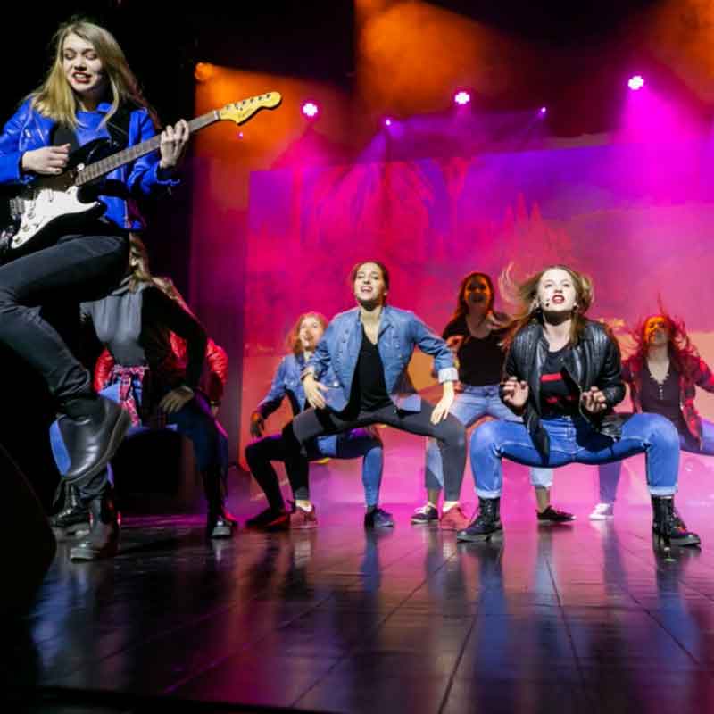  Na zdjęciu widoczna jest grupa kilku dziewcząt w tanecznym ruchu na scenie. Dziewczyna na pierwszym planie podskakuje z gitarą w rękach. Fot. Weronika Pawłowska - Ciągło