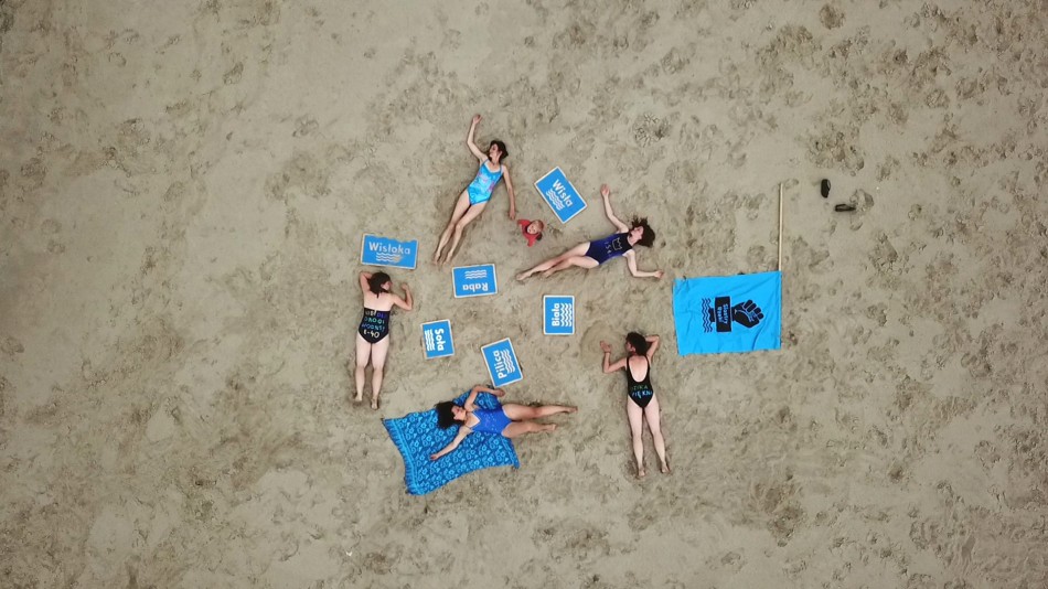 Zdjęcie przedstawia grupę kobiet leżących w kostiumach na plaży. Zdjęcie wykonane z lotu ptaka, od góry. Kobiety w kostiumach kąpielowych leżą na piasku, wokół porozrzucane niebieskie tabliczki z nazwami rzek Wisła, Raba , Biała, Wisłoka, Soła. Obok nich rzucona na piach niebieska flaga, z czarnym nadrukiem loga Siostry Rzeki, które przedstawi pięść zaciśniętą wynurzającą się z wody,. Wśród leżących kobiet stoi chłopiec patrzący w górę, widzimy jego wzrok.
