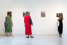 Na zdjęciu: Wystawa “Dla Puszczy” Michała Dąbrowskiego w Galerii Przy Kozach; trzy kobiety stoją tyłem do obiektywu, przyglądają się wiszącym na ścianie pracom. Fot. Służewski Dom Kultury