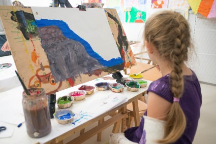 Na zdjęciu dziewczynka siedząca przed sztalugą. Przed dziewczynką jej praca oraz kolorowe farby. Dziewczynka maluje nimi obrazek. 