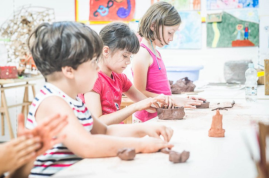 Zdjęcie ukazuje troje dzieci podczas warsztatów ceramicznych. Dzieci trzymają w dłoniach glinę. W tle wiszą rysunkowe prace plastyczne.