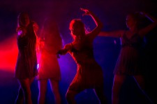 Zdjęcie przedstawia kilka tancerek w pozie tanecznej na scenie. Kolory światła scenicznego to czerwień oraz niebieski. 