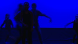Na zdjęciu widocznych jest kilka postaci w ruchu na scenie teatralnej. Widoczne kontury postaci oświetla niebieskie światło.