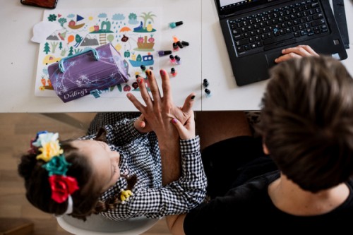 Na zdjęciu dziewczynka z Tatą, który pracuje na laptopie. Córka maluje mu paznokcie a na biurku widać kolorowe obrazki.