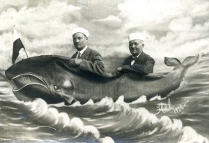 Dwóch mężczyzn pozuje do zdjęcia z namalowanym wielorybem. Mężczyźni ustawieni są przed dużą wymalowaną płachcie z wyciętymi dziurami symulującymi siedzenie na wielorybie. Fotografia w odcieniach szarości i sepii.