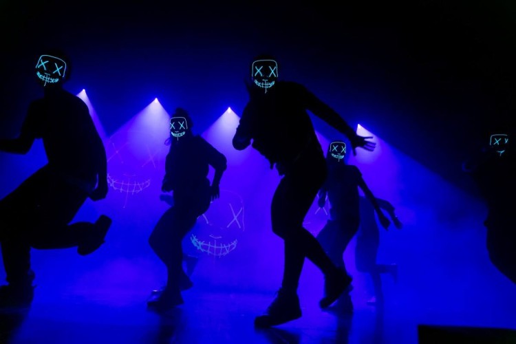 Na zdjęciu grupa tancerzy na scenie w ciemnym świetle. Przeważa kolor niebieski światła. Tancerze w maskach ze świecącymi oczami oraz ustami na twarzy.