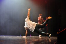Na zdjęciu tancerz Hip-Hop wykonujący akrobatyczną figurę. W tle widać chłopaka z mikrofonem. 