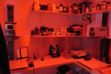 Na zdjęciu widoczne pomieszczenie ciemni. Czerwone światło. Półki z odczynnikami oraz sprzęt służący do wywoływania zdjęć. 