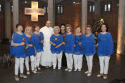 Na zdjęciu znajdują się członkinie zespołu Służewianki w towarzystwie księdza dominikanina. Wokalistki ubrane w niebieskie koszule i białe spodnie. Zdjęcie zrobione wewnątrz kościoła. 