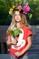 Na zdjęciu widoczna jest młoda dziewczynka w wianku z kwiatem w rękach. W tle widoczny jest amfiteatr SDK.