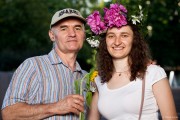 Na zdjęciu widoczna jest dwójka osób. Młoda kobieta oraz mężczyzna. Kobieta ma na głowie wianek, mężczyzna natomiast trzyma w ręku kwiat. W tle widać zieleń parku SDK.
