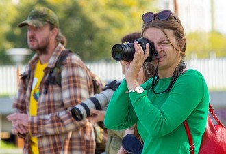 Na zdjęciu kobieta obserwująca ptaki za pomocą obiektywu fotograficznego próbując zrobić zdjęcie. W tle przewodnik projektu.