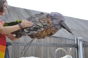 Uczestniczka warsztatów trzyma w dłoniach ptasią instalację plastyczną. W tle budynek SDK