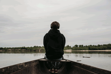 Kadrowanie centralne, linie zbieżne prowadzące do nastolatka siedzącego na dziobie łódki na rzece, chłopiec ma na uszach słuchawki i sfotografowany jest tyłem. Fot. Agnieszka Mocarska