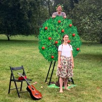 Fotografia: łąka, w tle park; na łące fragment dekoracji do przedstawienia "Bajka o drzewie" - owocująca jabłoń, przy niej aktor i aktorka. Obok drzewka krzesło, o które oparta jest gitara.