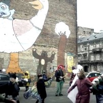 Kadr z teledysku grupy Wesołe Mazurki z Pragi: Fragment kamienicy z muralem przedstawiającym gęś; na placu przed kamienicą tańczące i grające na instrumentach dzieci z zespołu.