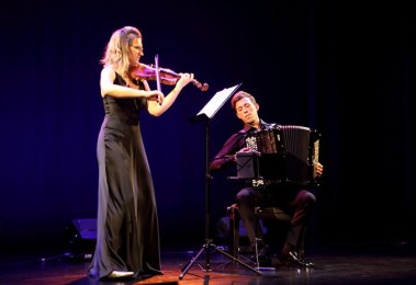 Na zdjęciu widać grający duet. Karolina Mikołajczyk stoi i gra skrzypcach. Ma czarną długą suknię i rozpuszczone blond włosy. Patrzy na stojak z nutami. Po prawej stronie siedzi Iwo Jedynecki i gra na akordeonie. Ubrany cały na czarno trzyma akordeon na kolanach. Scena jest oświetlona niebieskim światłem, w tle kotary.
