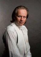 Na zdjęciu Paweł Kos-Nowicki w ujęciu do pasa, w białej koszuli na ciemnym jednorodnym tle.