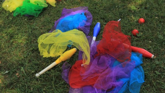 Zdjęcie przedstawia rozłożone na trawie rekwizyty do warsztatów cyrkowych - kolorowe chusty, kule i żonglerki