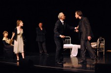 Zdjęcie wykonane w trakcie przedstawienia. Na zdjęciu aktorzy na scenie teatralnej. Mężczyzna na pierwszym rozmawia z innym mężczyzną. Obok stoi kobieta. W tle pozostali aktorzy. 