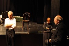 Zdjęcie wykonane w trakcie przedstawienia. Na zdjęciu aktorzy stojący przed sceną. Kobieta oraz mężczyzna stoją z założonymi rękami. 