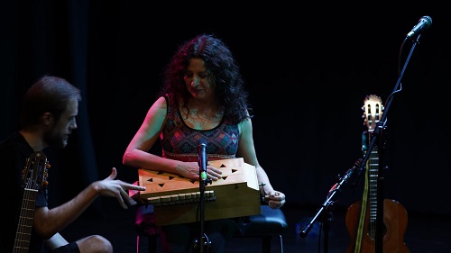Na zdjęciu widać Artystkę Annę Riveiro trzymająca na kolanach drewniany interument ludowy; słucha uwag dźwiękowca znadjującego się po lewej stronie zdjęcia. Ubrany jest na czarno i znajduje się poza głównym światłem. W tle gitary, statywy pikrofonowe, granatowe kotary sceniczne.