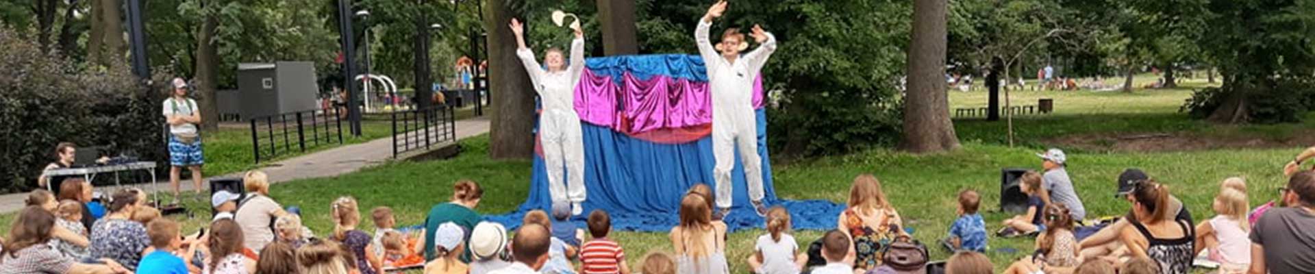 Kwadratowe zdjęcie. Grupa widzów siedząca w parku na kocach obserwuje aktorów na małej scenie na łące.