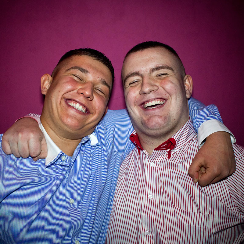Na grafice fragment zdjęcia, na którym dwóch młodych, uśmiechniętych mężczyzn obejmuje się za ramiona w braterskim uścisku.